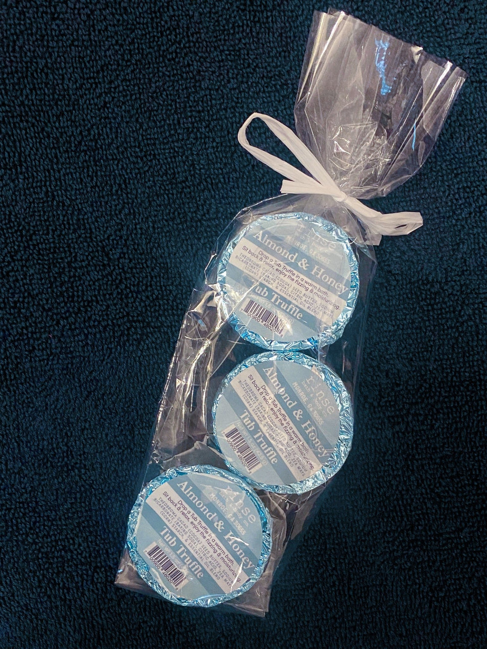 Almond & Honey Tub Truffle trio nestled in clear wrap on navy towel — soft blue foil labels in warm bath light