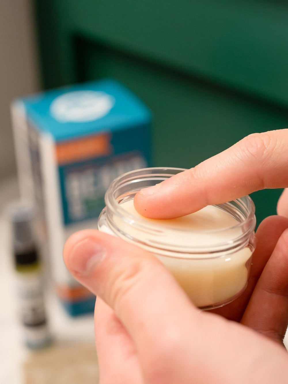 Finger pressing into pale Beard Balm on a marble vanity — soft bathroom light with a blue kit box blurred behind