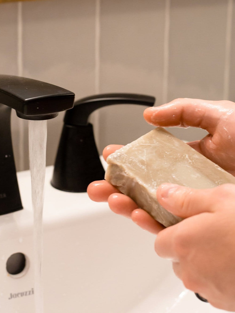 Hands working the Beard Bar into a lather at the bathroom sink — a cold-process face and beard soap with Dead Sea mud and bentonite clay. Rinse Bath & Body Co.