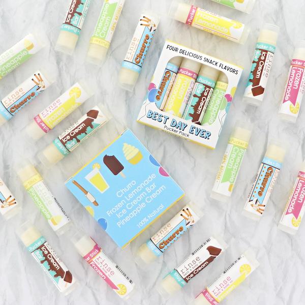 Best Day Ever lip balms scattered with their windowed pack on crinkled white cloth — treat icons in bright flat-lay light