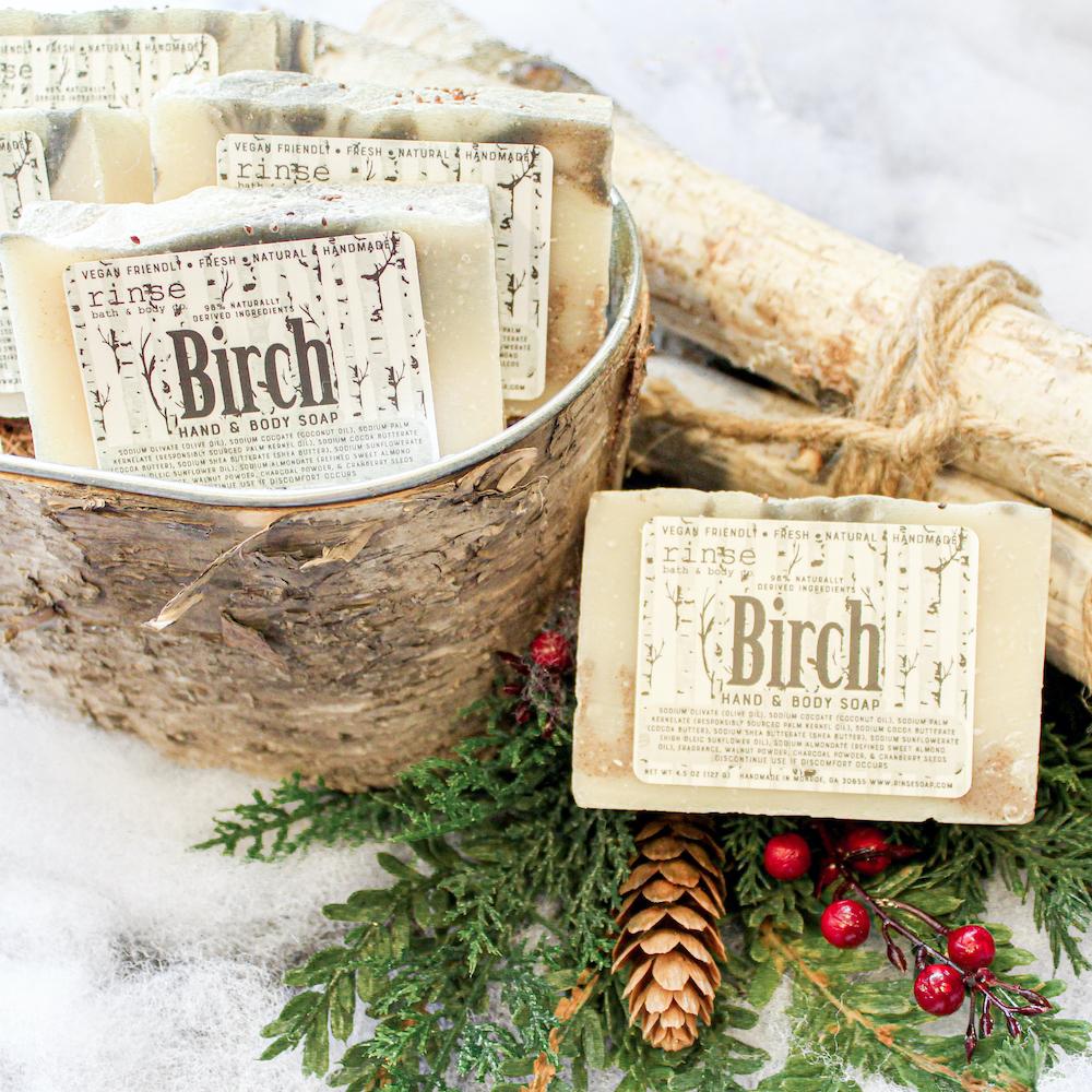 Birch-bark bucket of Birch soaps sits among logs and pinecone — red berries on snowy white with evergreens