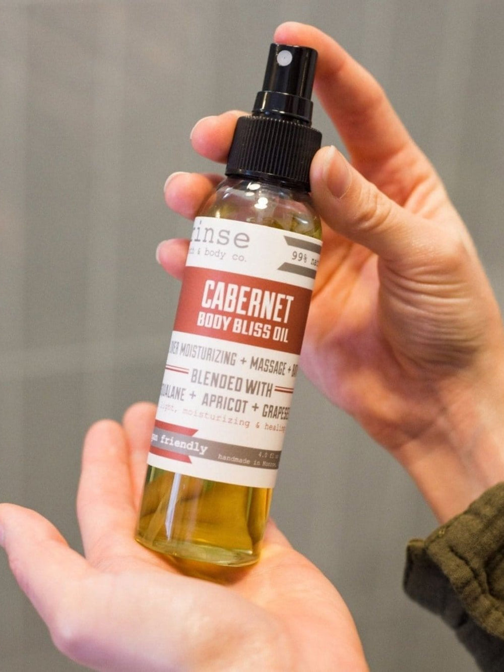 Hands cradling Cabernet Body Bliss Oil spray bottle — soft gray indoor light on the burgundy label