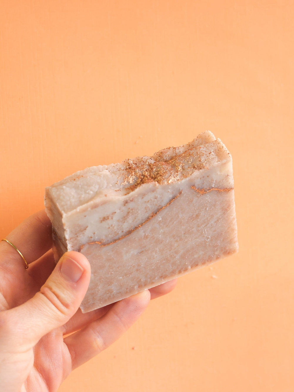 Hand holding Coco Kissed soap upright — creamy tan marble against vibrant apricot backdrop