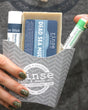 A hand holding the Dead Sea Mud fry-box gift set against concrete — Dead Sea Mud bar soap, Spearmint Lavender solid lotion stick and a Speppermint lip balm in the grey chevron carton. Rinse Bath & Body Co.