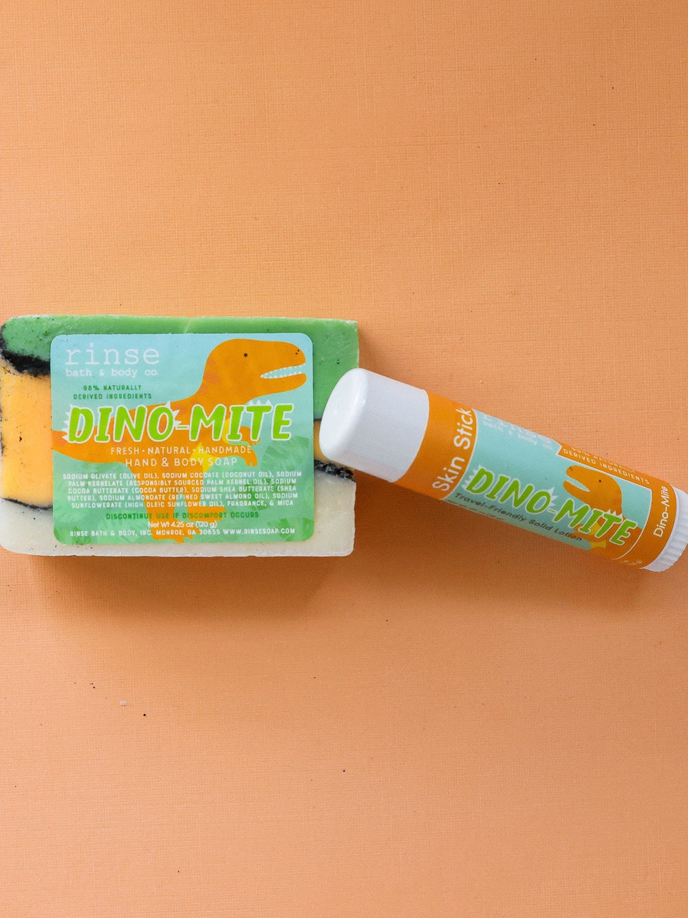 Dino-Mite Skin Stick beside layered green-black soap on vivid orange — matching dinosaur labels in flat-lay light
