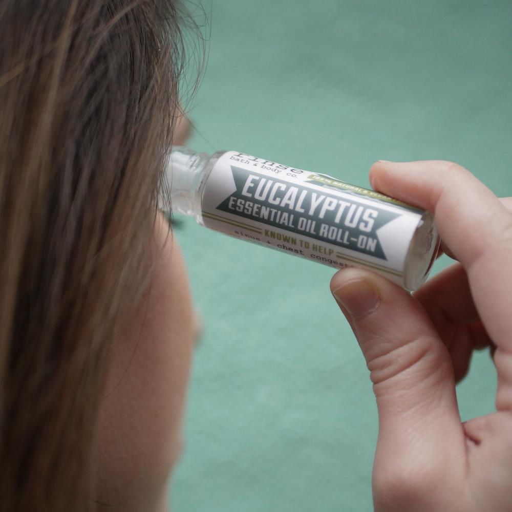 Hand applying Eucalyptus roll-on to a temple against muted teal — white-capped vial and soft daylight for cool herbaceous scent