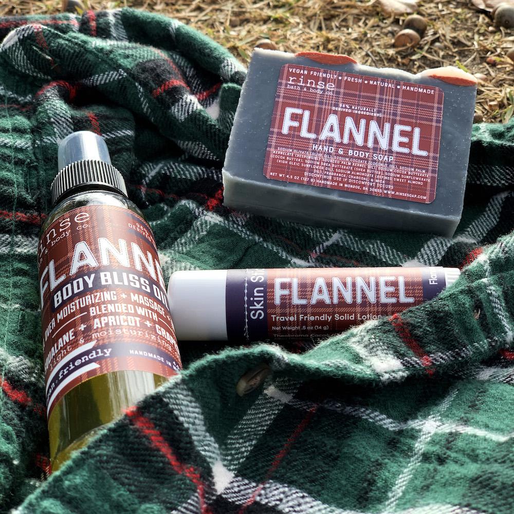 Flannel soap Body Bliss Oil and Skin Stick rest on a plaid shirt — dry autumn leaves and acorns outdoors