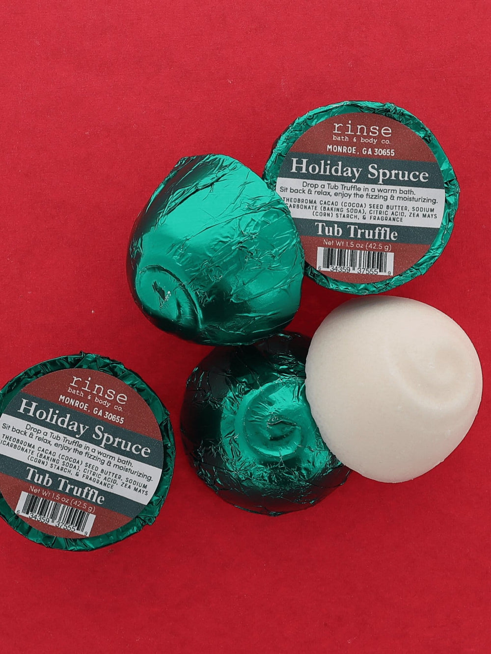 Two green foil-wrapped Holiday Spruce Tub Truffles and one unwrapped white dome on a deep red background — fresh evergreen for a winter bath. Rinse Bath & Body Co.