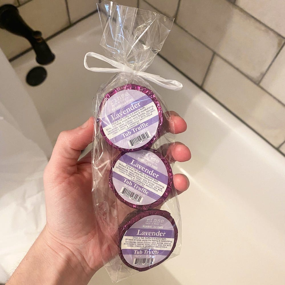Hand holding purple-foil Lavender Tub Truffle 3-pack — white subway tile bathroom behind