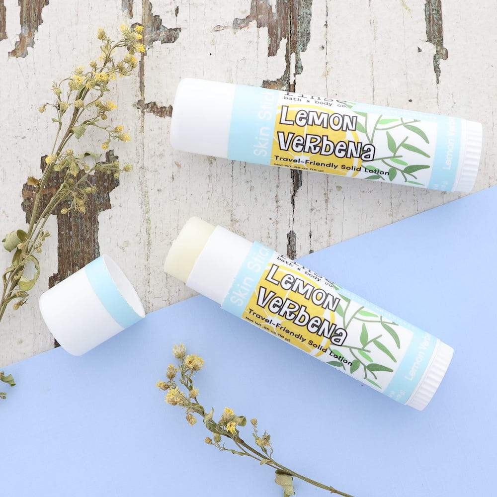 Open and capped Lemon Verbena Skin Sticks on distressed wood and light blue — yellow flower sprigs around the balm