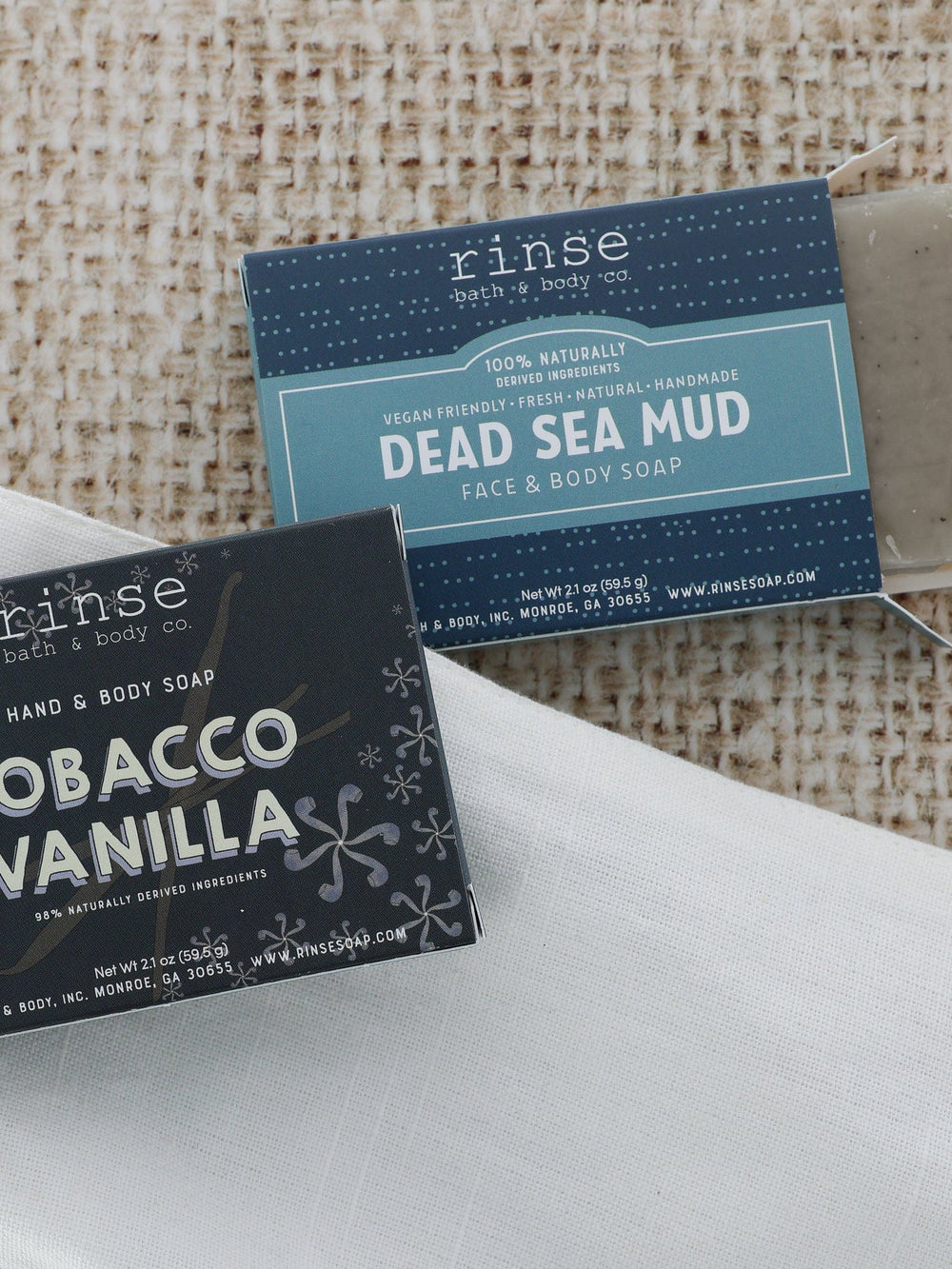 Tobacco Vanilla and Dead Sea Mud minis on linen and burlap — speckled gray soap sliding from its blue box