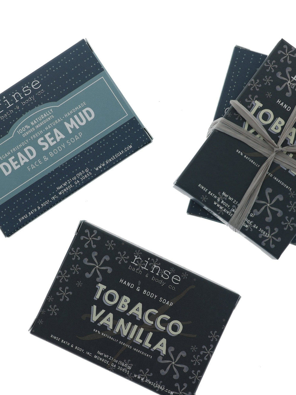 Dead Sea Mud teal box and Tobacco Vanilla charcoal wraps on bright white — gray raffia tying a gift stack