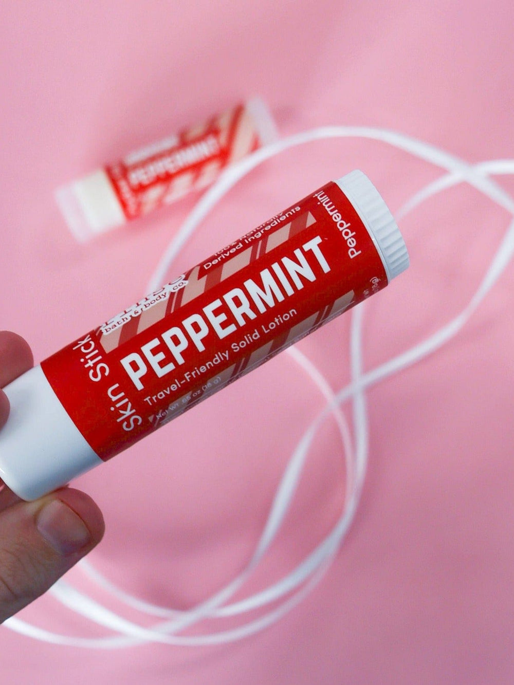 Hand holding Peppermint Skin Stick against bubblegum pink — white ribbons curling around the red tube