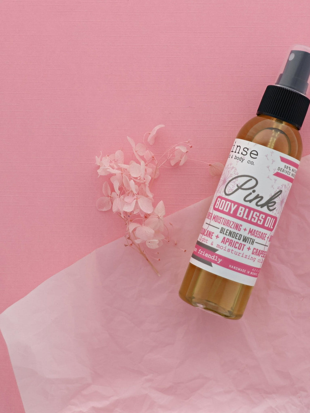 Pink Body Bliss Oil bottle beside delicate pink flowers on blush surface — spray bottle of golden oil in bright soft light