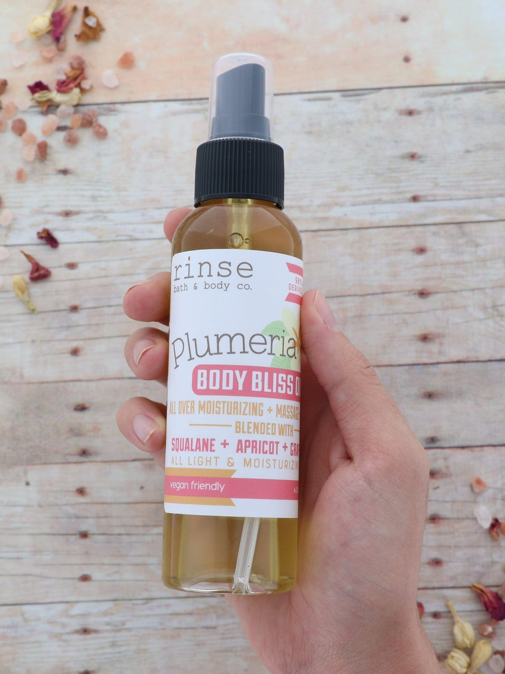 Hand presenting Plumeria Body Bliss Oil on weathered wood among petals and salt — golden spray bottle in soft daylight