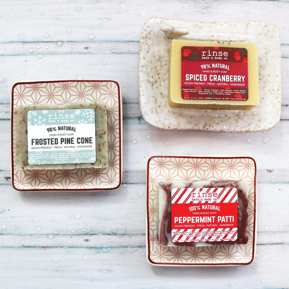Spiced Cranberry, Frosted Pine Cone, and Peppermint Patti bars on patterned dishes over droplet-dotted wood — festive trio in cool bathroom light