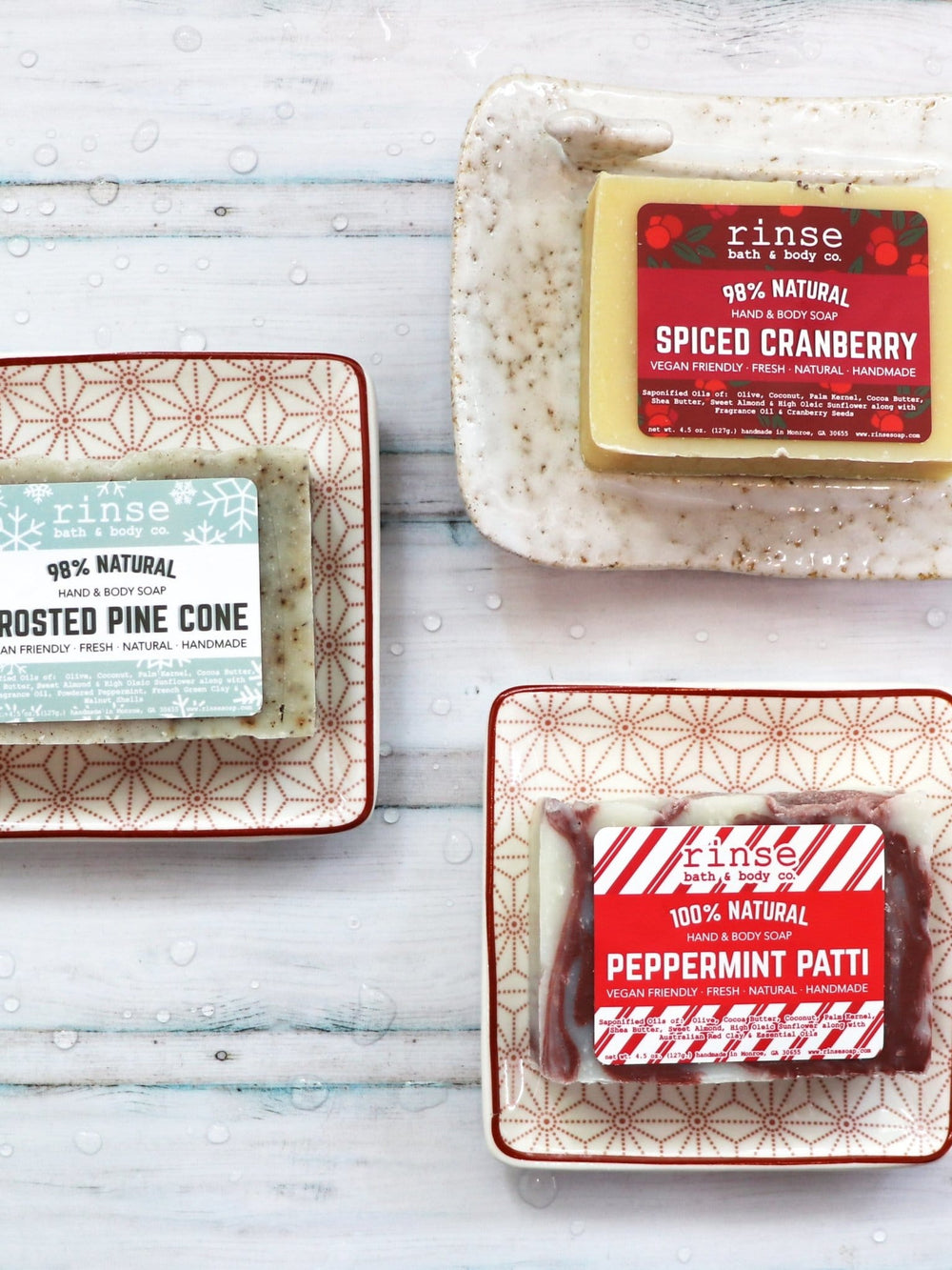 Spiced Cranberry, Frosted Pine Cone, and Peppermint Patti bars on patterned dishes over droplet-dotted wood — festive trio in cool bathroom light