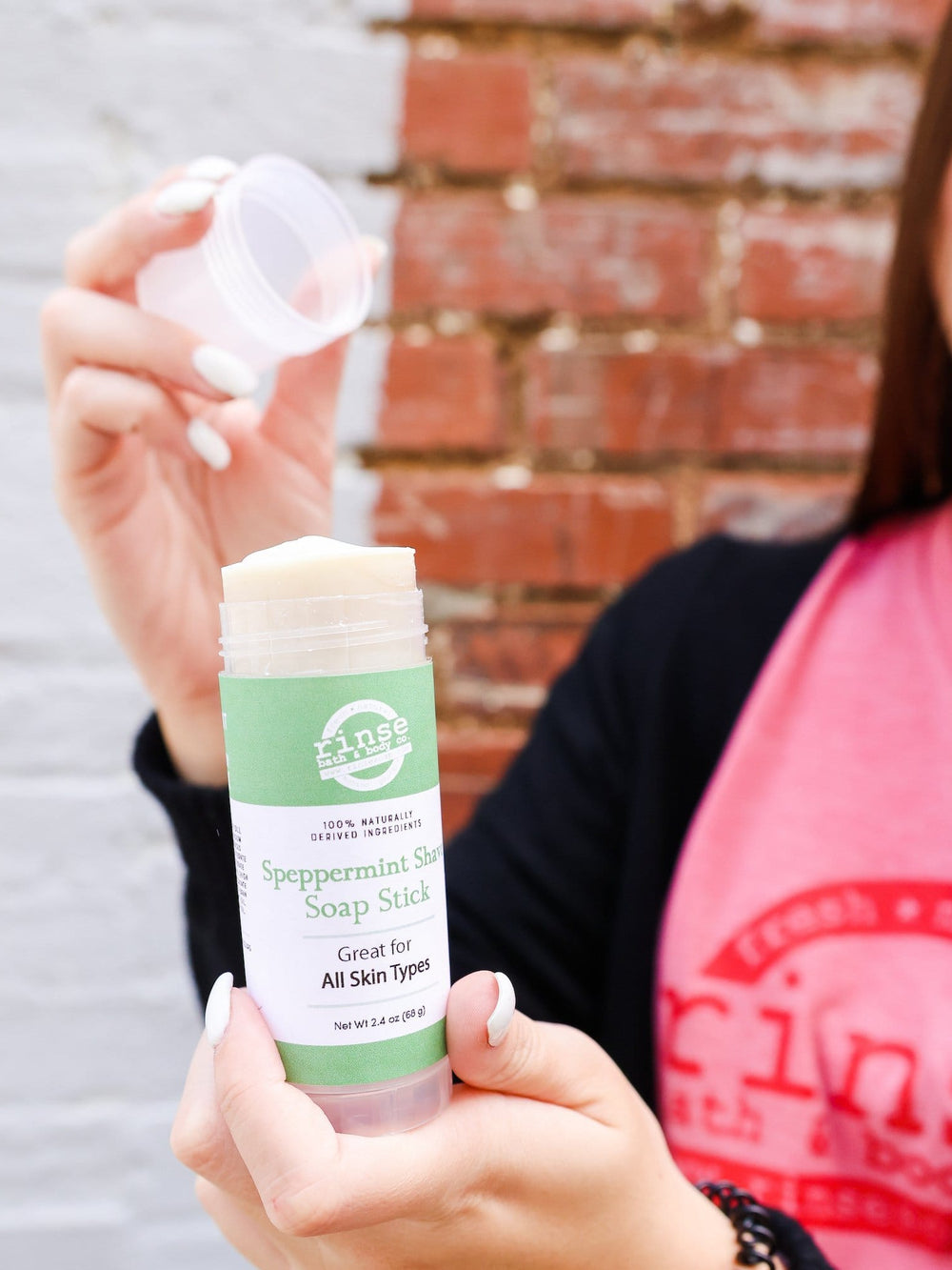 Hands uncapping Speppermint Shave Soap Stick before a half-painted brick wall — cream soap revealed in bright outdoor daylight