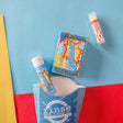 The Superhero kids gift set styled on blue, red and yellow colour-blocked paper — rainbow-swirl bar soap, solid lotion skin stick and lip balm beside the blue carton. Rinse Bath & Body Co.