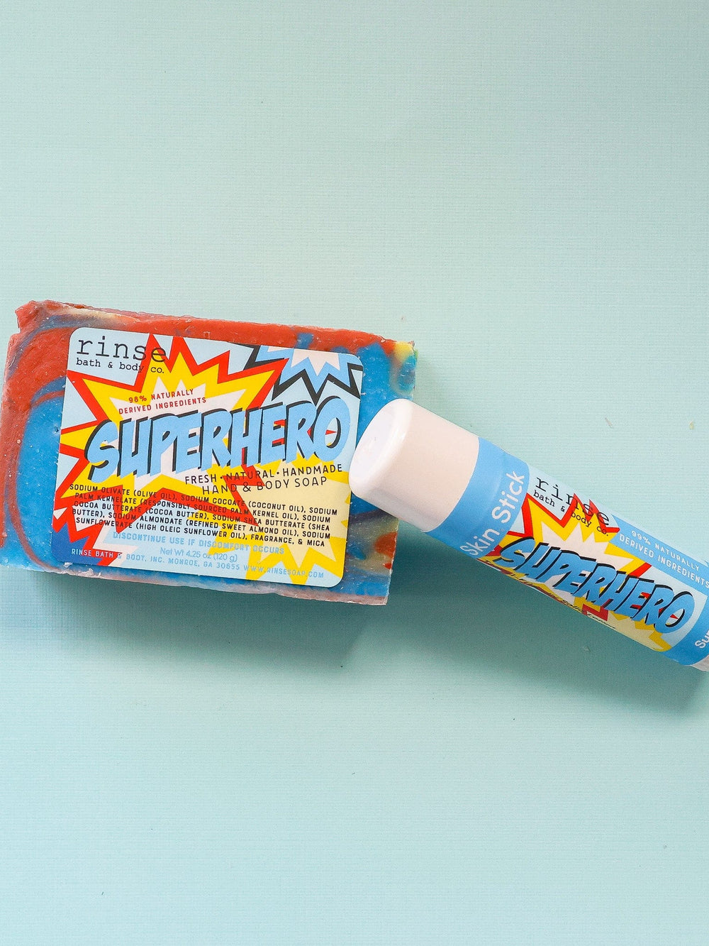 Superhero Skin Stick propped beside swirled blue and orange soap — seafoam table in bright clean light