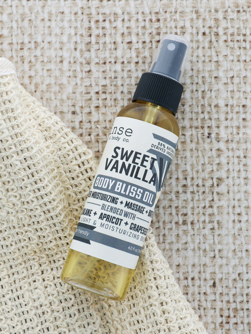 Sweet Vanilla Body Bliss Oil lying on a woven bath mitt — coarse cream fabric under the golden bottle
