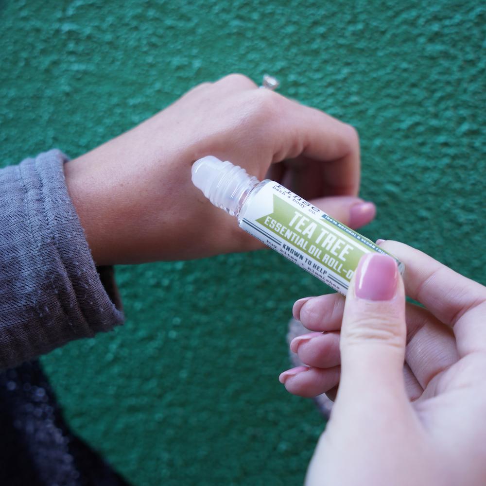 Pink-nailed hand showing oil sheen on the wrist beside open Tea Tree roll-on — textured teal wall behind