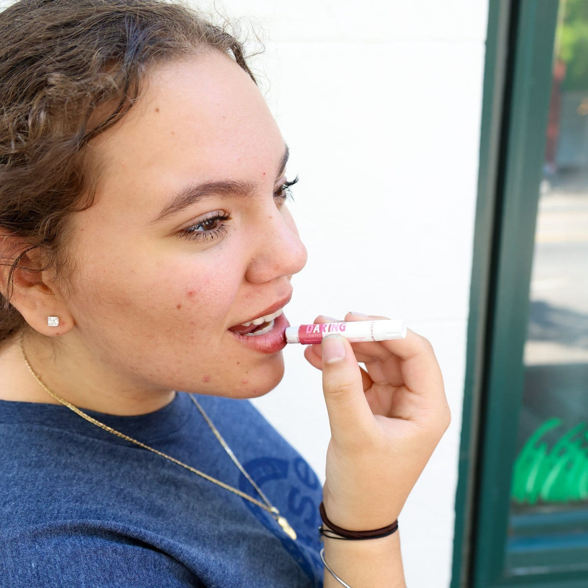 A woman in scrubs applying the Daring tinted lip balm in front of a glass door. Rinse Bath & Body Co.