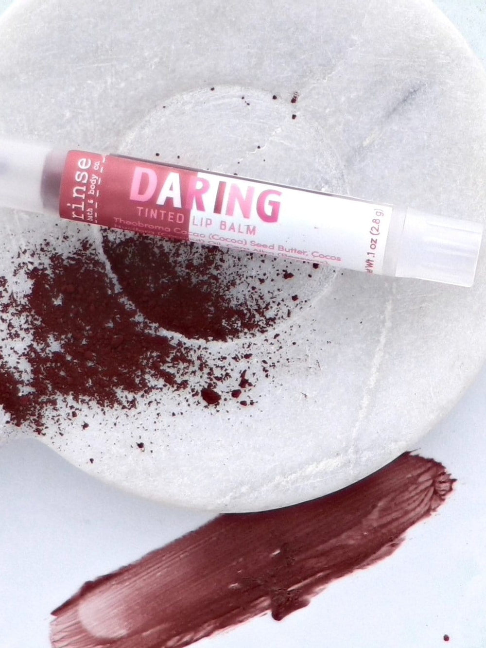 Daring tinted lip balm resting on white marble beside maroon pigment and a waxy swatch — cool clean texture light