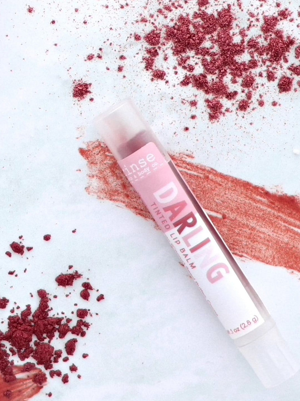 Darling tinted lip balm tube on a shimmery coral swatch with scattered pigment — textured stone flat lay