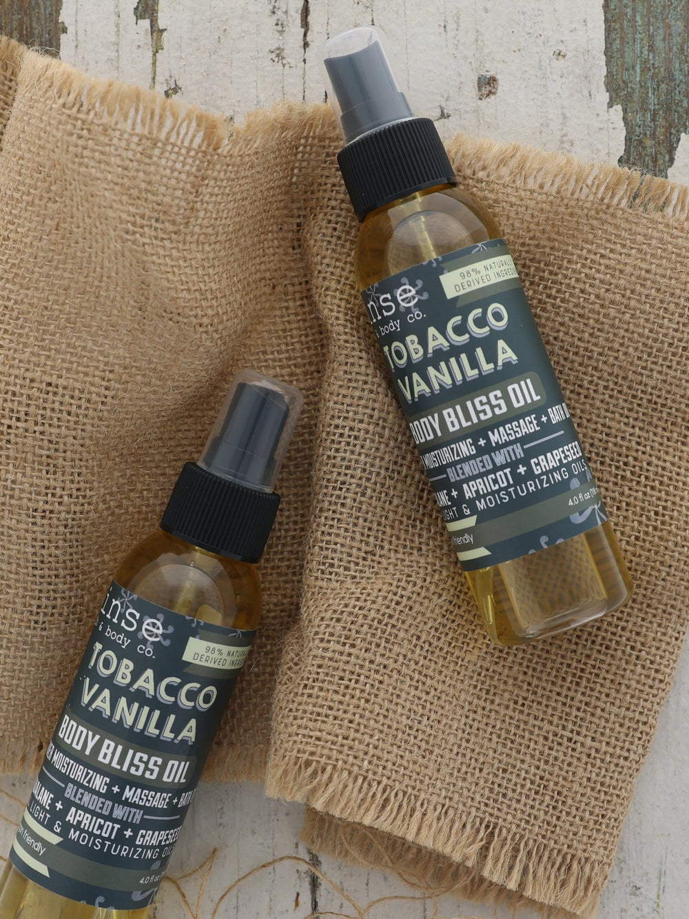 Two Tobacco Vanilla oil bottles on light burlap over weathered painted wood — rustic overhead flat lay