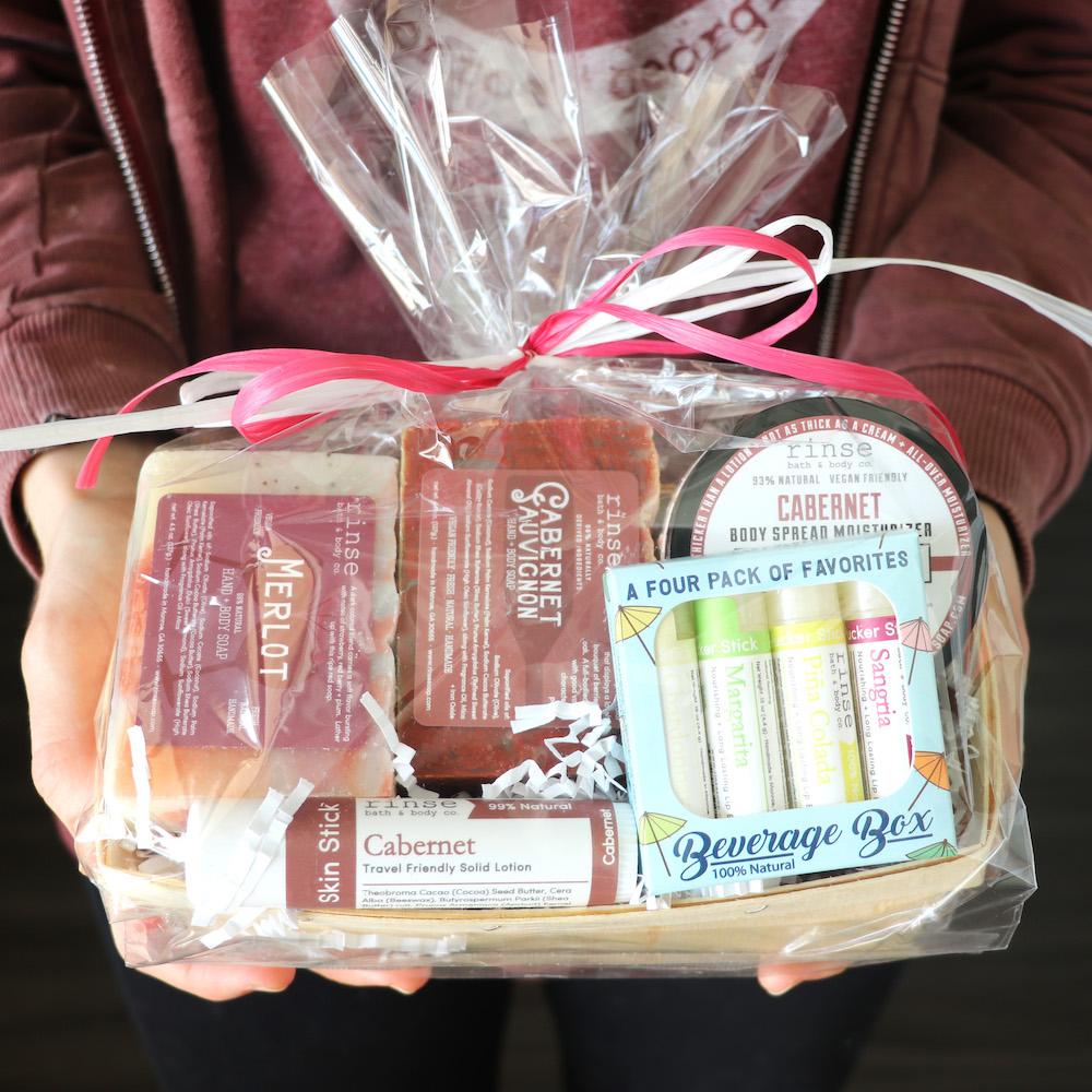 Hands in a maroon hoodie hold the Wine Gift Basket — cellophane, pink bow, wine-scented set inside.